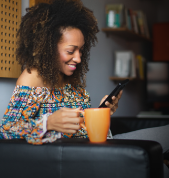 A black woman sitting on her couch using her cell phone while drinking coffee