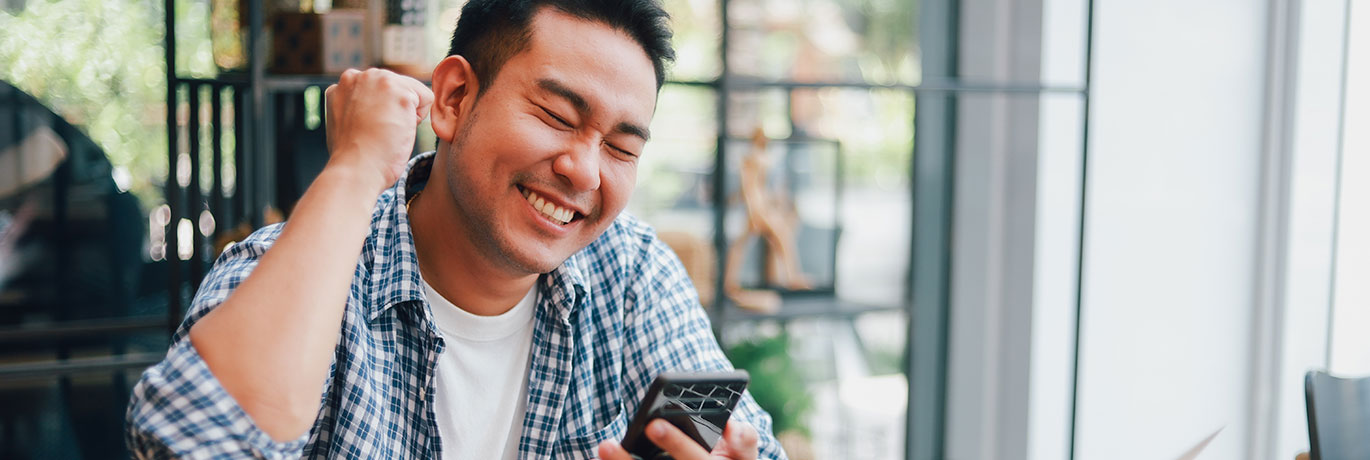 Young Asian man sitting outside celebrating with his phone