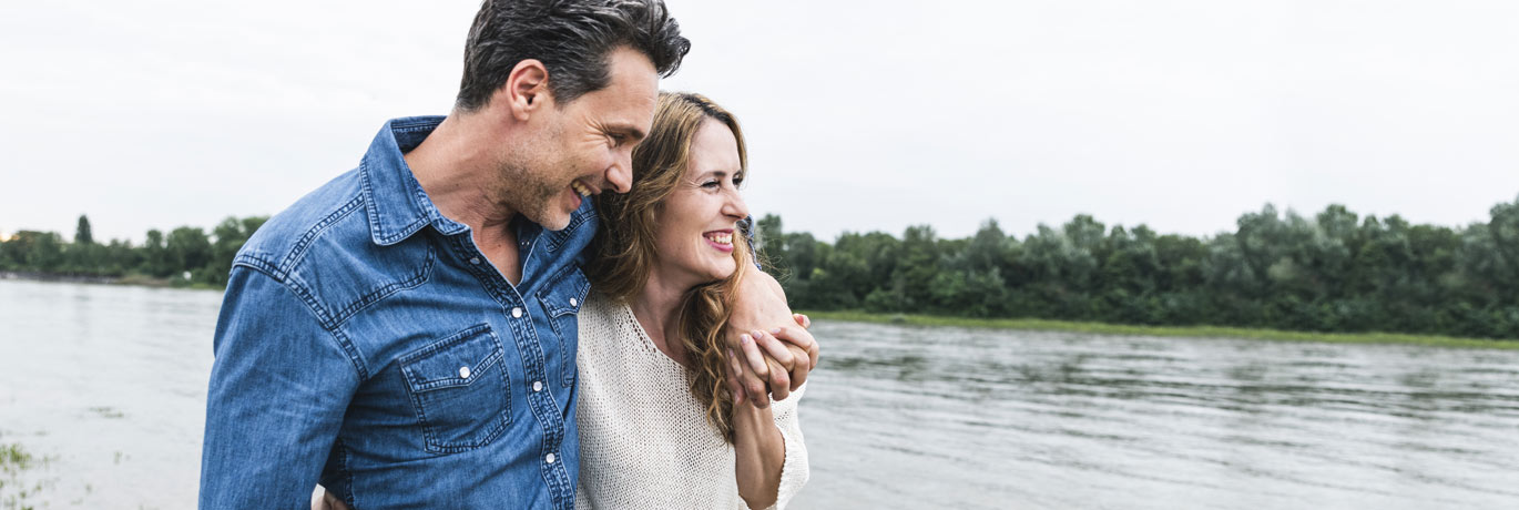 A man and woman with arms around each other smiling while walking along a lake