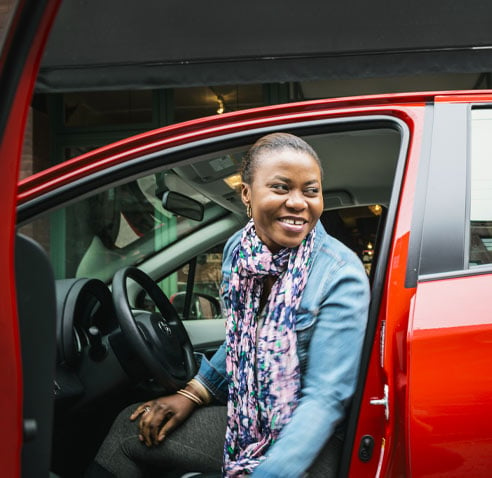 A black woman sitting in her car with the door open, smiling at someone outside.