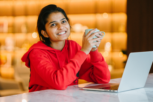 An Indian woman sitting infront of her laptop holding a coffee cup