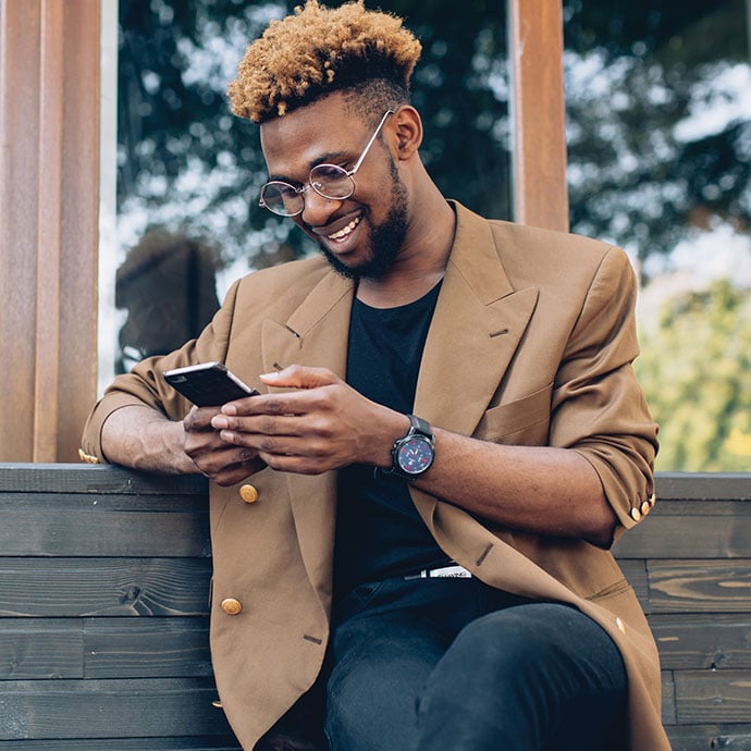 A young well-dressed black man sitting on a bench outside using his cell phone.