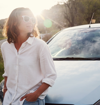 A young women leaning on the hood of her car smiling in the sun.